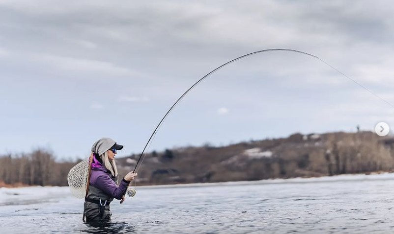 image by @thebugparade of Riversmith brand ambassador Paula Shearer fly fishing in Canada with a nice bend on her rod and signature purple jacket