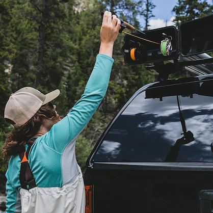 Women wearing fishing waders reaching up to slide a fishing rod into an open fishing pole roof rack. It is mounted to the top of a black pickup truck with a matching black bed cap