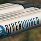 Close up of the river quiver logo on the side of the silver metal of the 4-banger river quiver’s rod casing. The logo says “river” in black text and “quiver” in blue text with a matching blue image of fishing bait hitting the water. The background is blurred out but looks vaguely like vegetation. 
